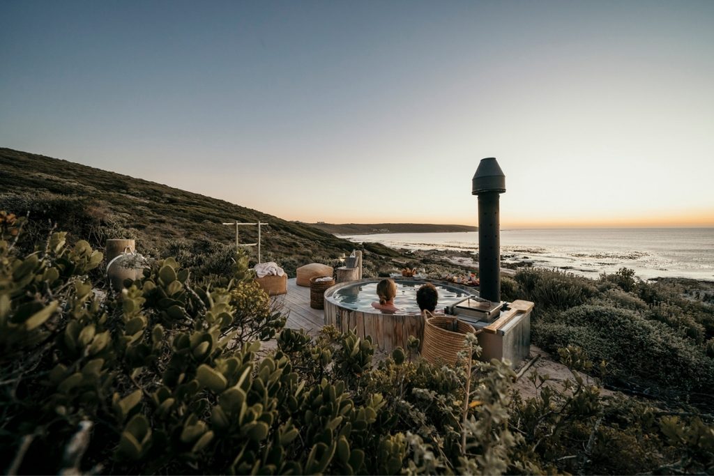 Couple relaxing in a Buitengewoon wood-fired hot tub overlooking the ocean at Pure Sea in Romansbaai